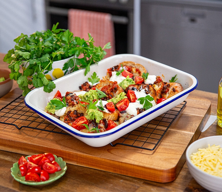 A plate of crispy wholemeal roll nachos topped with melted cheese, diced tomatoes, avocado, and black beans, served on a rustic wooden table.