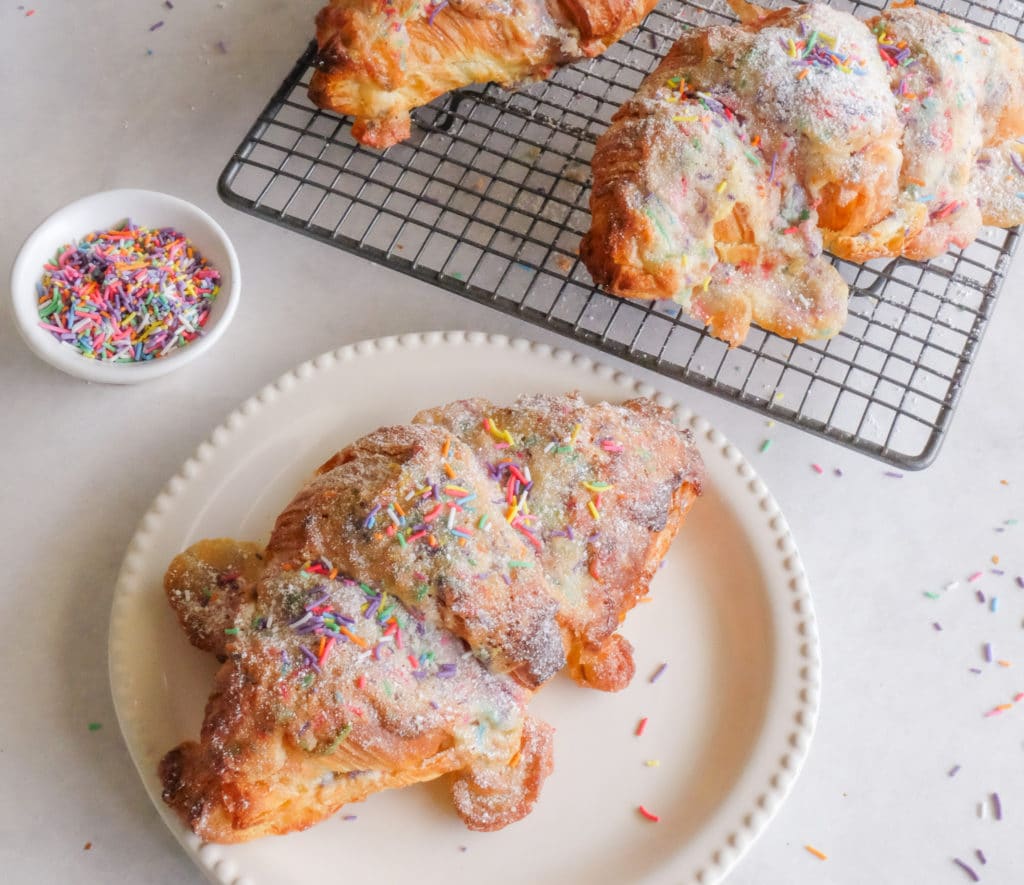 A golden, flaky birthday cake croissant filled with a sweet cake-flavored filling and topped with colorful sprinkles.