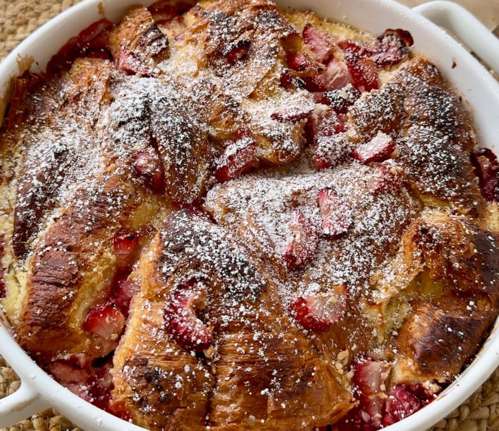 A warm, golden-baked Strawberry Cheesecake Croissant Pudding topped with fresh strawberries and a dusting of icing sugar.