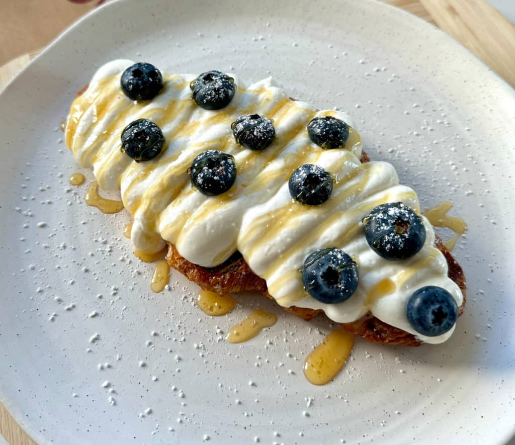 A golden croissant waffle (croffle) topped with creamy ricotta, fresh blueberries, and a drizzle of honey, served on a rustic plate.